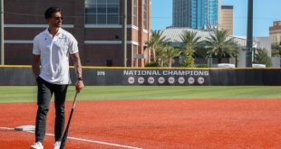 Man in a white polo shirt and dark pants standing on a baseball field holding a bat, with a National Champions sign on the outfield fence and city buildings behind him.