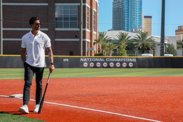 Man in a white polo shirt and dark pants standing on a baseball field holding a bat, with a National Champions sign on the outfield fence and city buildings behind him.