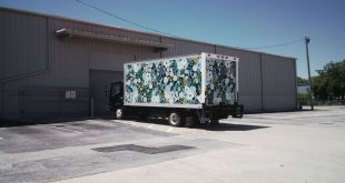 A delivery truck with a floral wrap parked at a warehouse loading dock under a clear blue sky.