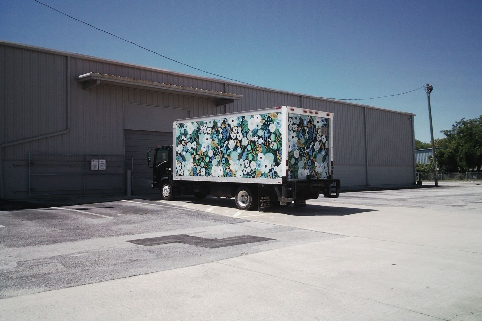 A delivery truck with a floral wrap parked at a warehouse loading dock under a clear blue sky
